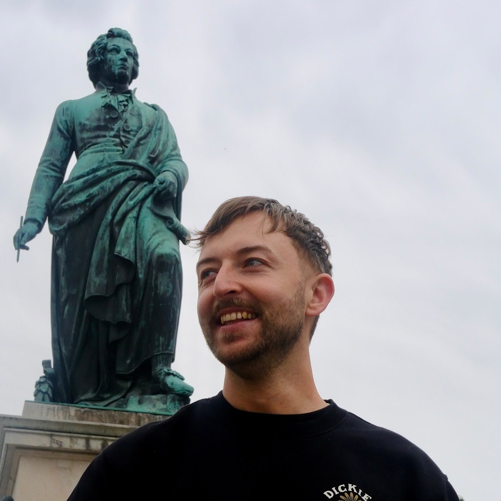 Breucko lacht und blickt zur Seite, im Hintergrund eine Bronzestatue auf einem Platz.