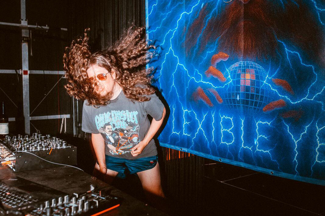 Flexible Yeah wirbelt mit wehenden Locken an den DJ-Reglern, im Hintergrund ein Banner mit Discokugel-Motiv und Blitzen.